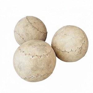 May include: Three vintage-style beige leather balls with visible stitching. One ball has the "deBeer" logo. The balls are arranged in a group, suggesting a set or collection. The balls appear to be used for a sport or game.