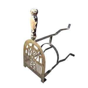 May include: A vintage fireplace tool featuring a brass front plate with an intricate cut-out leaf design, a vertical handle with distressed black and white paint, and a dark metal frame with two angled prongs extending from the back for support.