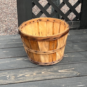 May include: A rustic, wooden basket with a round shape and a wide opening. The basket is made of light-colored wood with darker wooden bands around the top and middle. Metal wire handles are attached to the sides. The basket sits on a dark gray wooden surface.