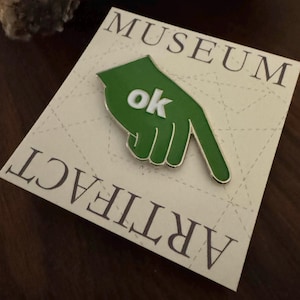 May include: Green enamel pin shaped like a hand pointing down with the word "ok" in white letters. The pin is on a card that says "Museum Artifact" at the bottom and "Museum" at the top.