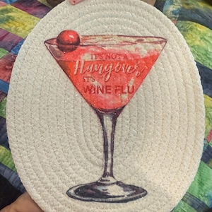 May include: Round, off-white coaster with a glittery, woven texture. The coaster features a red cocktail illustration with the text "It's Not a Hangover, It's Wine Flu." A small, pink tag with the word "love" is attached.