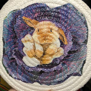 May include: A white rope trivet with a colorful image of a brown bunny rabbit sitting inside a purple cabbage.
