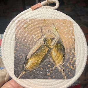 May include: A white rope coaster with a printed image of two champagne flutes with gold glitter. The flutes are raised in a toast.