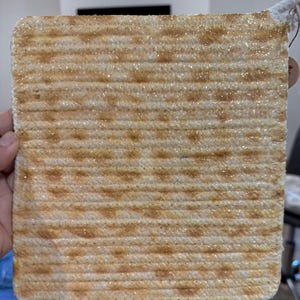 May include: A square, woven coaster with a light tan and brown color scheme. The coaster has a textured surface with a repeating pattern of small, darker brown spots. It is held by a hand, with a tag attached to the top.