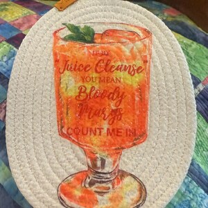 May include: A round, woven coaster with a printed image of a Bloody Mary cocktail in a glass. The text reads "If by Juice Cleanse You Mean Bloody Marys Count Me In". The coaster has a loop for hanging.