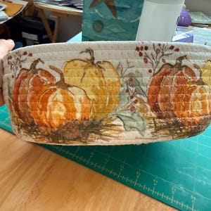 May include: A round, off-white rope basket featuring a printed design of orange and yellow pumpkins, green leaves, and red berries. The basket is constructed from coiled fabric rope, showcasing a textured surface with a seasonal harvest theme.