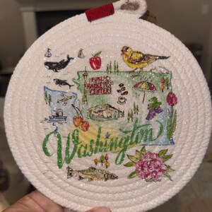 May include: Round, white rope coaster with a hand-painted map of Washington State. The map includes illustrations of a whale, apple, bird, and the words "Public Market Center" and "Washington". A decorative item.