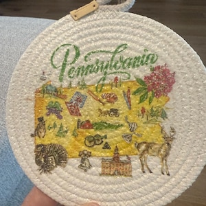 May include: Round, woven coaster with a map of Pennsylvania. The coaster features a yellow background with green text that says "Pennsylvania". The map includes illustrations of landmarks and symbols, with a deer and a turkey.