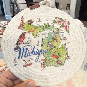May include: Round, white woven trivet featuring a map of Michigan with various illustrations. The map includes a lighthouse, bear, deer, flowers, and the word "Michigan" in blue. A small leather tag with the word "love" is attached.