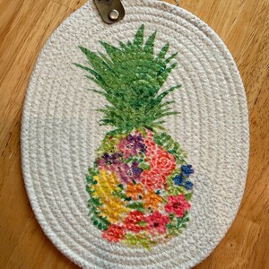 May include: An oval-shaped white coiled rope trivet featuring a central pineapple graphic. The pineapple has a green leafy top and a body filled with a colorful floral pattern in shades of pink, yellow, orange, purple, and blue. A small leather tag with a metal rivet is attached to the top.