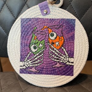 May include: Round white coaster with a purple center featuring two cartoon cocktails held by skeleton hands. One cocktail is green with an eye, the other orange with a tooth. The coaster has a tag that says "Hand made with love".