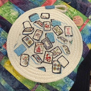 May include: A round, off-white trivet with a rope-like texture, adorned with hand-painted Mahjong tiles in various colors. The trivet has a blue loop for hanging. A small tag is attached to the trivet.