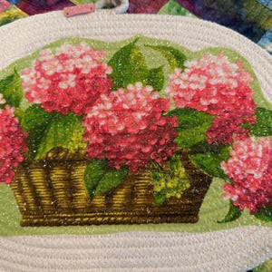May include: Round, white woven placemat with a floral design. The design features a basket of pink and red hydrangeas with green leaves. The placemat has a small, pink tag with the word "LUV" on it.