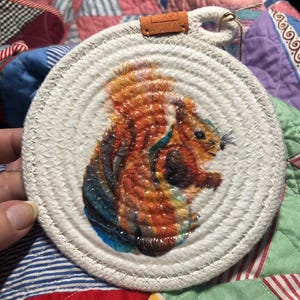 May include: A round, white, rope-wrapped coaster with a painted image of a red squirrel. The squirrel is depicted in a colorful, abstract style.