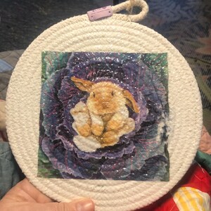 May include: A white round woven trivet with a colorful image of a brown bunny rabbit sitting inside a purple cabbage.