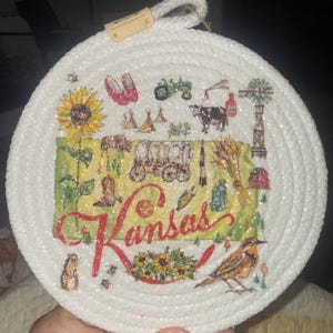 May include: A round, white rope trivet with a colorful Kansas-themed design. The design includes a sunflower, a tractor, a cow, a windmill, and the word "Kansas" in red script. The trivet has a loop for hanging.