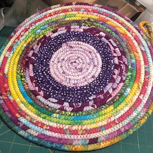 May include: A colorful round braided fabric trivet with a purple and white center. The trivet is made of multiple layers of fabric strips sewn together in a circular pattern.