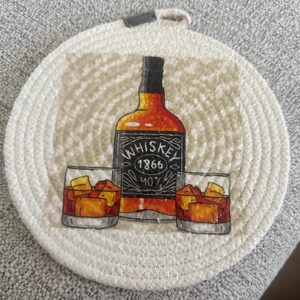 May include: Round white coaster with a printed design featuring a whiskey bottle and two glasses of whiskey with ice. The bottle has the word "Whiskey" and the numbers "1866" and "40%" printed on it. The coaster has a loop for hanging.