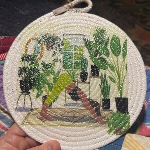 May include: A round white crocheted trivet with a printed image of a person doing yoga in a room with plants. The trivet has a rope handle.