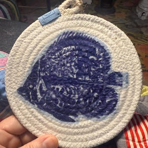May include: A white rope coaster with a blue and white fish design. The coaster is round and has a rope edge.