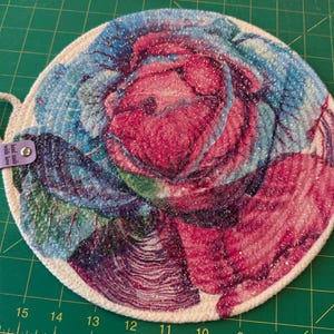 May include: Round, braided trivet with a floral design. The center features a large, pink and red flower with blue and green accents. The trivet has a loop for hanging and a small tag that says "hand made with love".