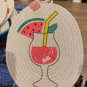 May include: Round, off-white coaster with a glittery surface and a cocktail design. The cocktail features a watermelon slice, yellow straw, and green leaf. A small tag is attached with a string.