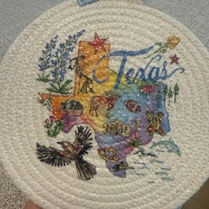 May include: Round, woven coaster featuring a colorful map of Texas with iconic imagery and the word "Texas". A small blue tag with "love" is attached. The coaster has a rope loop for hanging.
