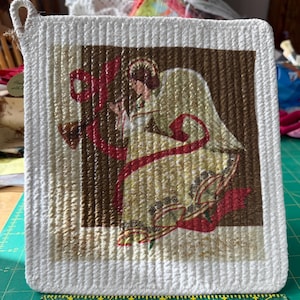 May include: Square white and beige pot holder with a loop for hanging. The center features a brown background with an angel illustration in gold, red, and beige. The angel is holding a red ribbon.