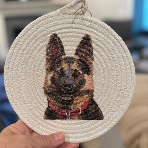 May include: Round, off-white woven coaster featuring a detailed portrait of a German Shepherd dog. The dog has brown and black fur, a red collar, and a focused expression. A loop of natural twine is attached for hanging.