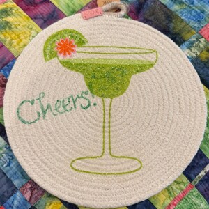 May include: Round, off-white coaster with a margarita design. The margarita glass is green with glitter, and a lime wedge and flower accent the drink. The word "Cheers!" is written in green script. A small tag says "love".