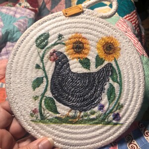May include: Round, off-white woven trivet with a painted hen design. The hen is black and white with a red comb, surrounded by sunflowers and greenery. A small leather tag with the word "HANDMADE" is attached.