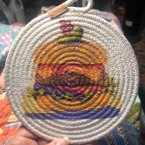 May include: A white round woven pot holder with a colorful image of a hamburger on it. The hamburger has a red tomato, green lettuce, and a yellow bun.