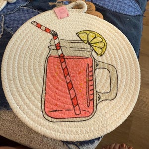 May include: A round, cream-colored coaster with a hand-drawn illustration of a pink drink in a mason jar with a red and white striped straw and a lemon slice. The coaster is made of woven material.