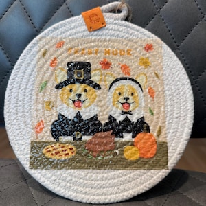 May include: Round, woven trivet with a Thanksgiving theme. Features two corgi dogs dressed as pilgrims, with the words "FEAST MODE" above them. Includes images of fall leaves, pie, and a pumpkin.