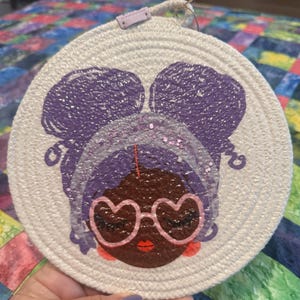May include: Round, off-white coaster with a graphic of a Black woman's face. She has purple hair, heart-shaped glasses, and red lips. The coaster is made of woven material and has a small loop for hanging.