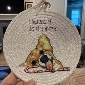 May include: Round, white woven coaster with a dog illustration and the text "I licked it, so it's mine." The dog is brown, black, and white, with a candy cane in its mouth. A loop of rope is attached for hanging.