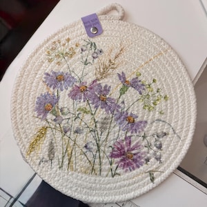 May include: A round, off-white coiled rope trivet featuring a printed floral design with purple daisies, green stems, and golden wheat stalks. A small, light purple fabric tag at the top is printed with the text "hand made with love".