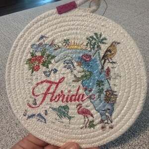 May include: Round, white woven coaster featuring a colorful map of Florida with the word "Florida" in red script. The map includes illustrations of a flamingo, dolphin, and palm trees. A small tag with the word "love" is attached.
