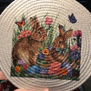 May include: A white rope trivet with a printed design of two brown rabbits surrounded by colorful flowers and butterflies. The rabbits are holding Easter eggs.