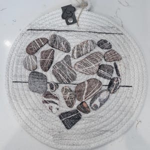May include: A round, white woven coaster with a heart-shaped arrangement of stones. The stones are various shades of gray, brown, and white, with striped patterns. A black leather tag with a heart shape is attached to the top.