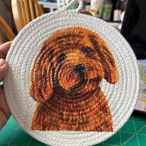 May include: A round, off-white woven trivet with a painted portrait of a brown dog. The dog has a black nose and dark eyes. A loop is attached for hanging. The trivet is approximately 8 inches in diameter.