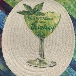 May include: Round, off-white woven trivet with a green cocktail design. The cocktail glass has the words "NO WORKING DURING Drinking HOURS" printed on it. The trivet has a loop for hanging.
