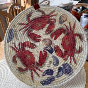 May include: Round, woven trivet featuring a nautical design with red crabs and blue and gray seashells. The trivet has a cream-colored background and a loop for hanging. The word "Crab" is visible on the trivet.