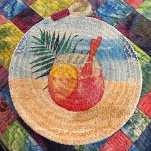 May include: Round, hand-painted coaster featuring a tropical cocktail with a slice of orange and two red straws. The coaster has a beach scene with palm leaves and the word "love" on a small tag. The coaster is made of a woven material.