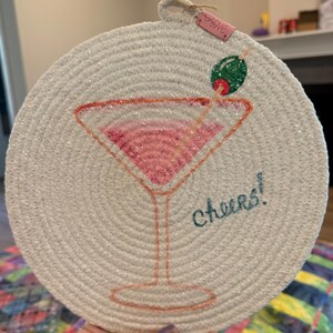 May include: Round, off-white coaster with a stitched design of a pink cocktail in a martini glass. The coaster has the word "Cheers!" in teal and a green olive with a red toothpick. A small tag with the word "Love" is attached.