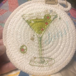 May include: A white crocheted coaster with a green martini glass and olives printed on it. The coaster has a brown tag that says "Handmade".