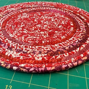 May include: A round, braided trivet in shades of red, burgundy, and white. The trivet is made of fabric strips sewn together in a spiral pattern. It has a loop for hanging and a small tag attached.