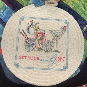 May include: Round, white woven coaster with a blue border and the phrase "GET YOUR mahj ON." The coaster features a watercolor illustration of cocktails and playing cards. A blue loop is attached for hanging.