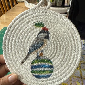 May include: Round white woven coaster with a painted bird perched on a striped ornament. The bird has a holly berry and leaf detail on its head. The coaster has a loop for hanging.