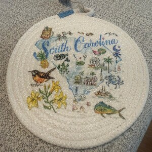 May include: Round, white woven trivet featuring a map of South Carolina with illustrations of local landmarks and wildlife. The words "South Carolina" are written in blue script. A small blue tag with the word "love" is attached.